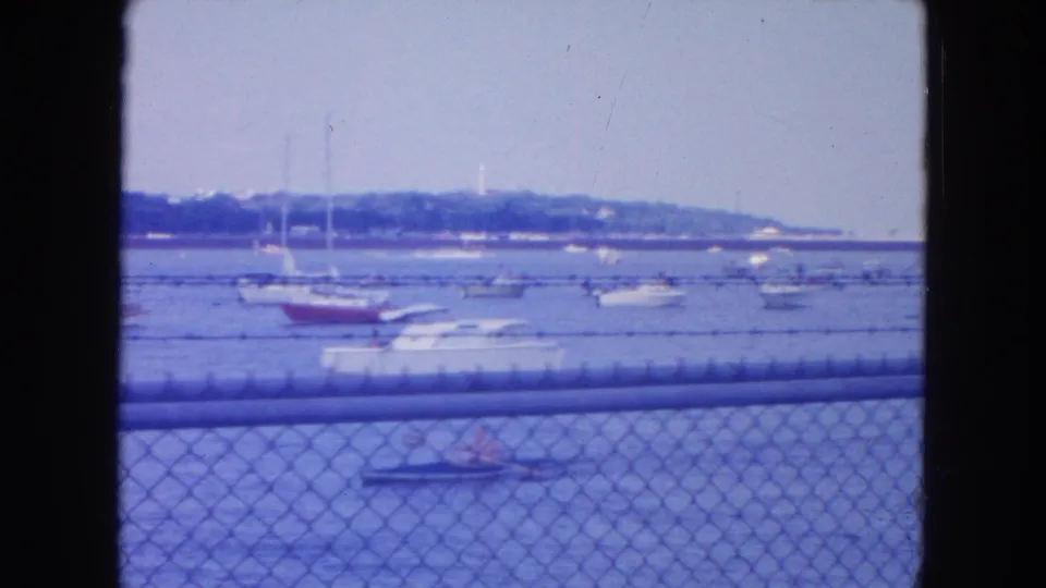 1954 New York City waterfront view with small boats, sailboat masts, and a chain-link fence in soft 8mm color.