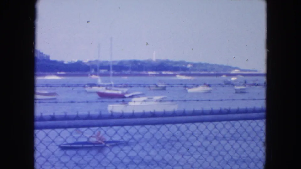 1954 New York City 8mm frame showing moored boats behind a fence and a small foreground boat with a distant person aboard.