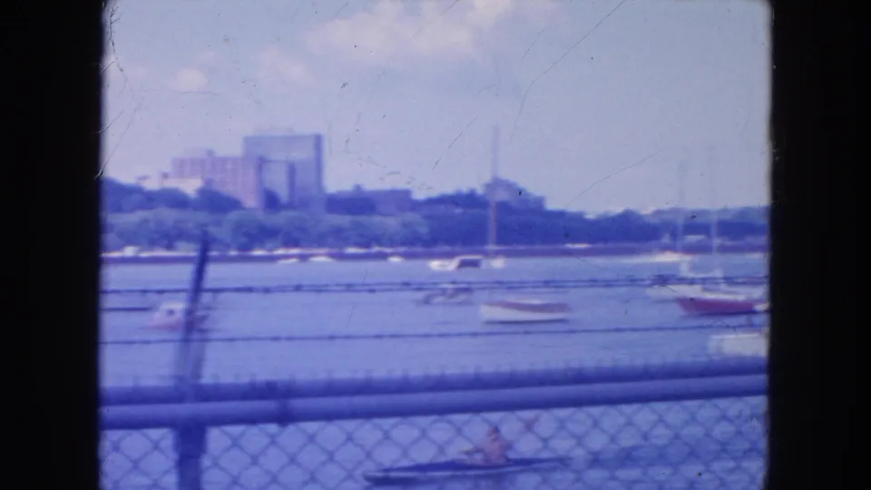 1954 New York City waterfront frame with small boats, a foreground paddled boat, fence rails, and distant buildings across the water.