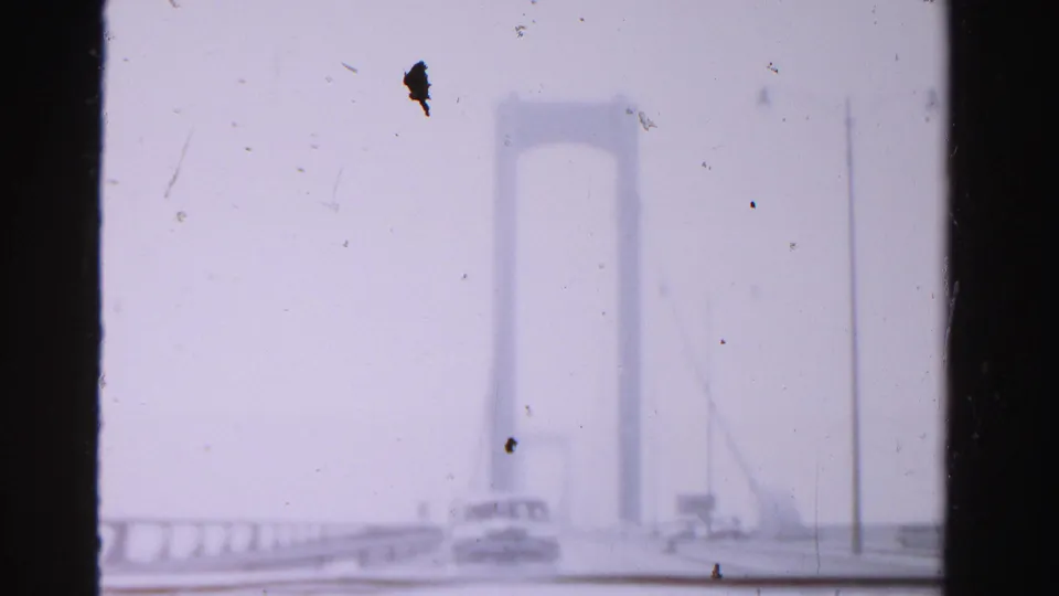 1955 Dade County home-movie frame of a car on a suspension bridge, with tower, cables, and roadway railing softened by haze.