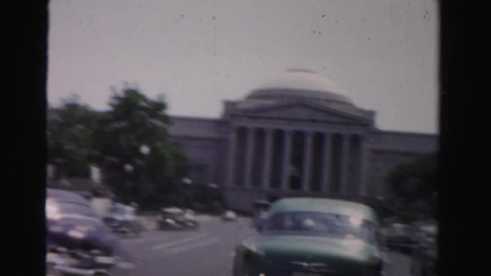 1955 Dade County home-movie frame with a domed civic-looking building, trees, and mid-century cars on the road.