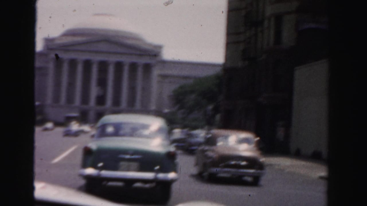 Green and brown 1950s cars pass a white columned building on a city street.