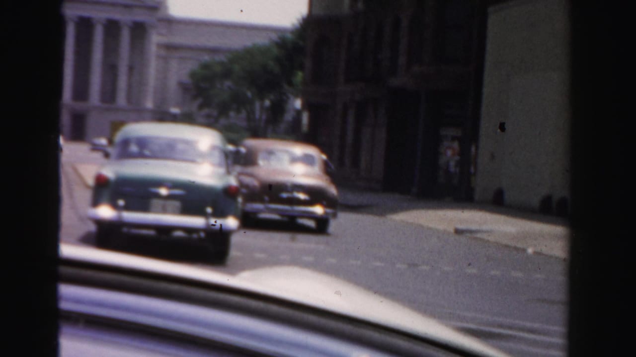 Drive-by 1955 Dade County view of two 1950s cars on a street near a white columned public-style building and darker storefronts.