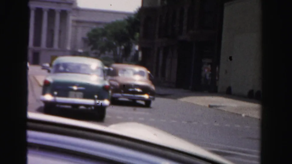 Drive-by 1955 Dade County view of two 1950s cars on a street near a white columned public-style building and darker storefronts.