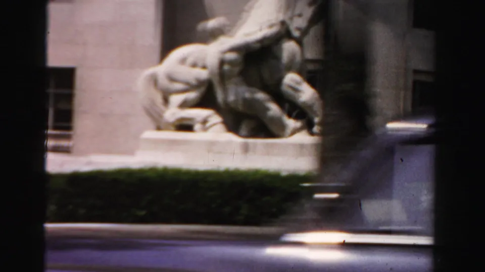 1955 Dade County frame showing a large pale stone horse-and-rider statue near a building facade, hedge, and passing car-window edge.