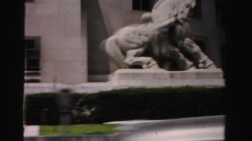 Soft 8mm drive-by view of a pale horse-and-rider statue in Dade County, with building wall, hedge, lawn, and window framing visible.