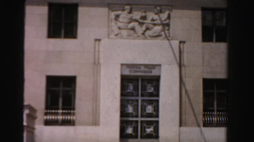 1950s 8mm frame of a pale civic building facade with relief sculpture, dark windows, and gridded entrance doors.