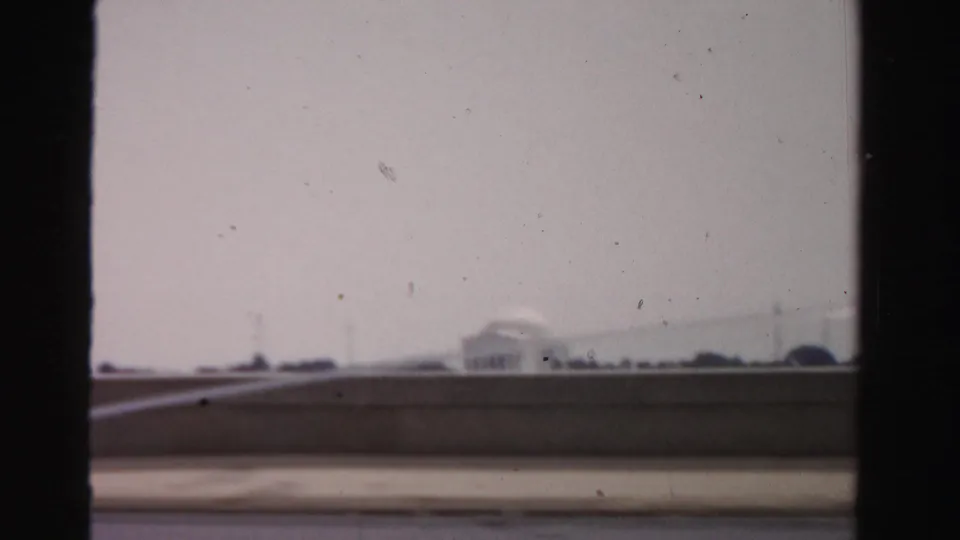 1950s 8mm frame showing a distant memorial-like structure beyond a low wall, filmed under a washed pale sky.