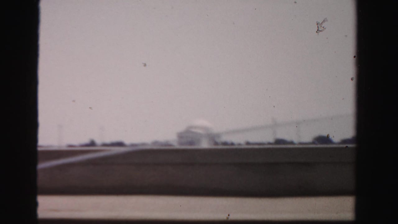 1950s 8mm frame looking through dark vertical framing toward water and a distant domed memorial-like building across the basin.