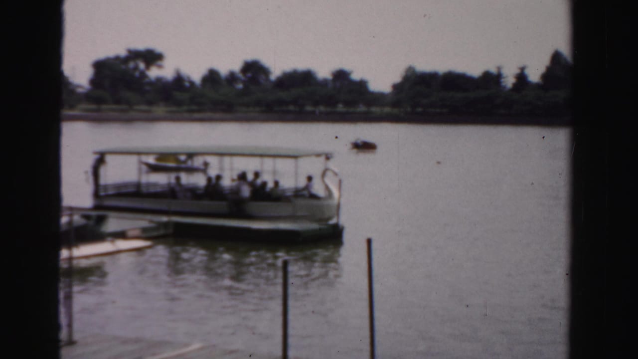 A covered sightseeing boat carrying visible passengers sits on calm water near a tree-lined shore in faded 8mm footage.