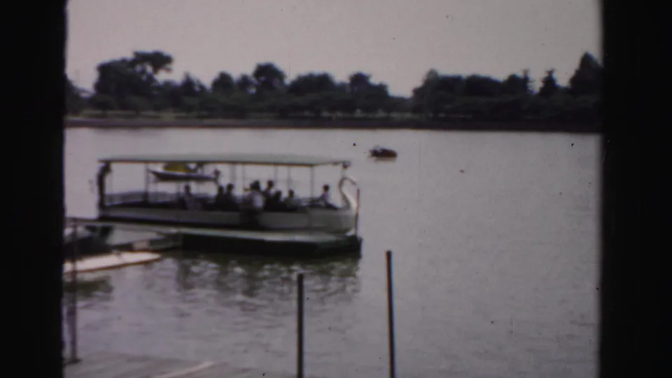 A covered sightseeing boat carrying visible passengers sits on calm water near a tree-lined shore in faded 8mm footage.