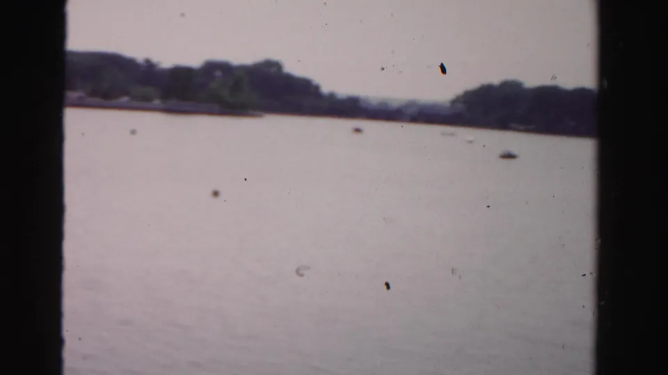 Wide, soft 8mm view over calm water toward a tree-lined shore, with the memorial no longer centered in frame.