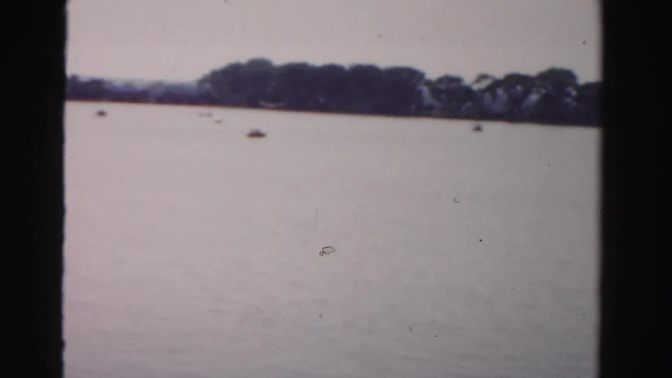 Soft archival frame of calm water, distant trees, and small boats on a broad waterside landscape.