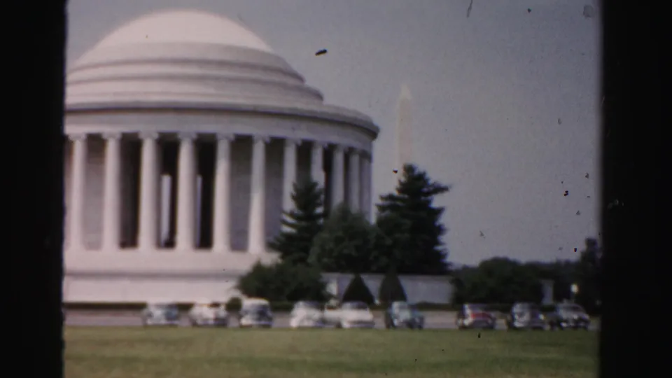 1955 8mm frame showing the Jefferson Memorial, parked cars, lawn, trees, and a faint Washington Monument in the background.