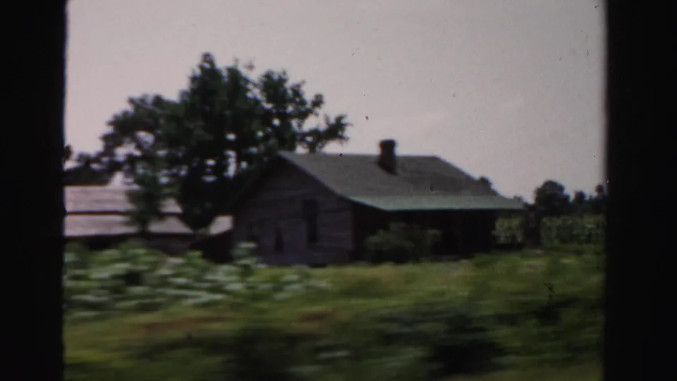 Dade County farm scene from 1955 with a weathered farmhouse, green crops, trees, and roadside grass captured on 8mm film.