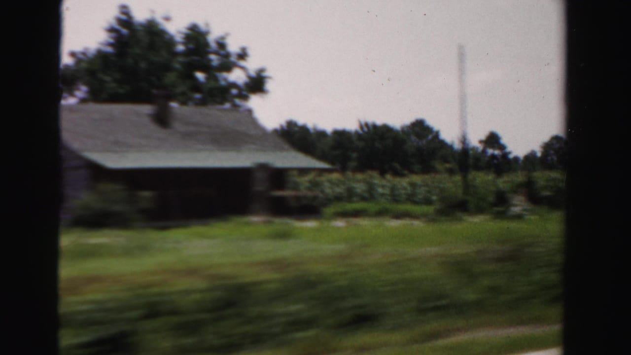 Blurred roadside view of a rural house, trees, grass, and fields from a moving car.