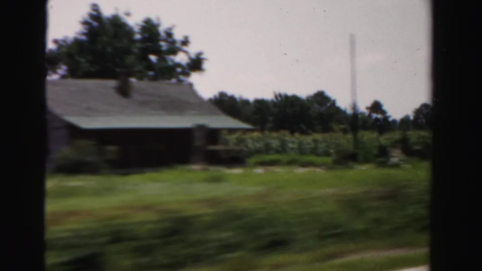 1955 Dade County roadside view of a weathered rural house, trees, grass, and open fields filmed through a moving-car frame.