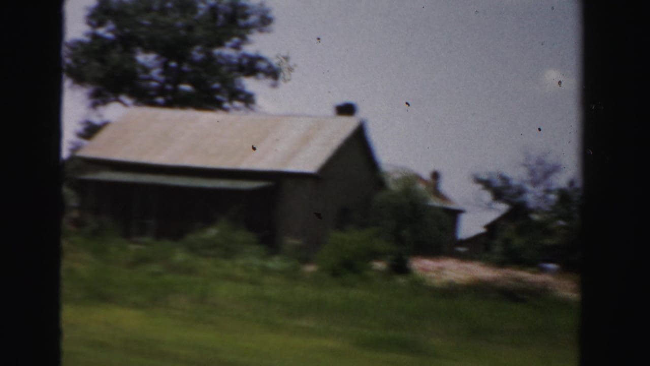 Rural building with a light roof, trees, grass, and a yard seen from a moving vehicle.