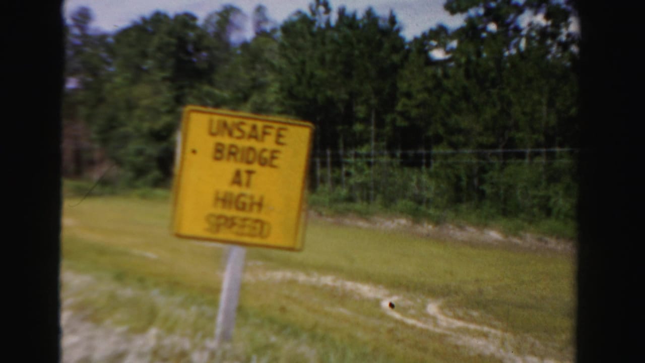 Yellow roadside sign warning of an unsafe bridge at high speed.