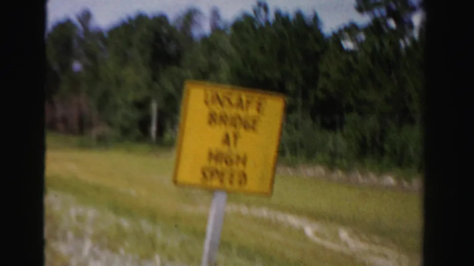 Motion-blurred 1955 road view of a yellow unsafe-bridge warning sign against grass and trees.