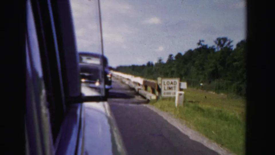 1955 Dade County passenger-window view showing a classic car ahead, concrete bridge barriers, and a load-limit sign.