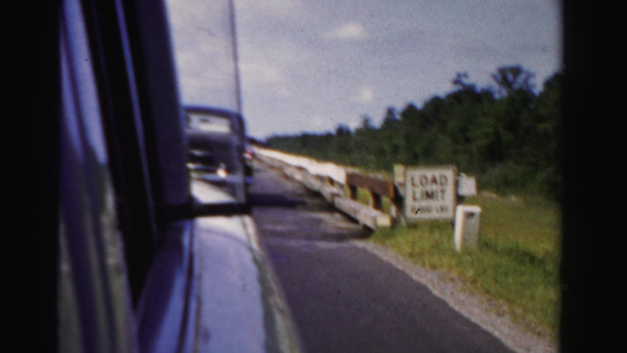 1955 Dade County passenger-window view showing a paved road, white guardrail, trees, sky, and a vintage car ahead.
