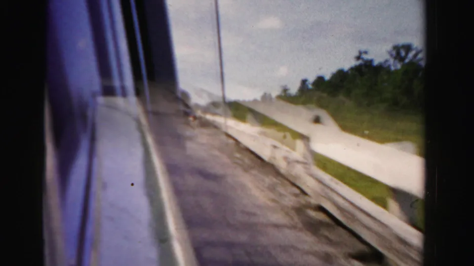 1955 home-movie frame from a moving car in Dade County, with the window edge, guardrail, road shoulder, grass, trees, and bright sky.