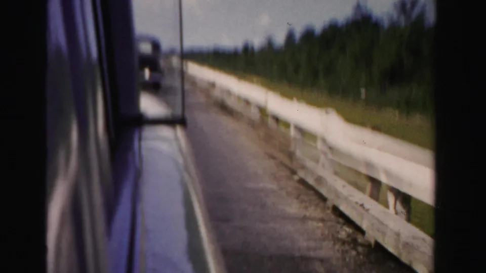 Dade County road view from inside a moving car, showing a white guardrail, paved lane, side mirror, trees, and sky.