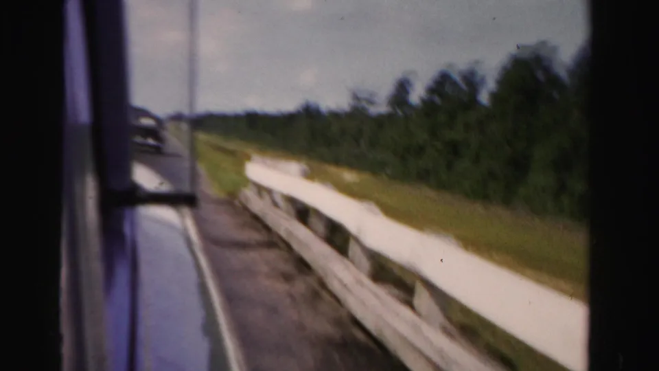 The 8mm frame shows a blurred 1955 Dade County roadside with white guardrail, green trees, grass, and the vehicle edge at left.