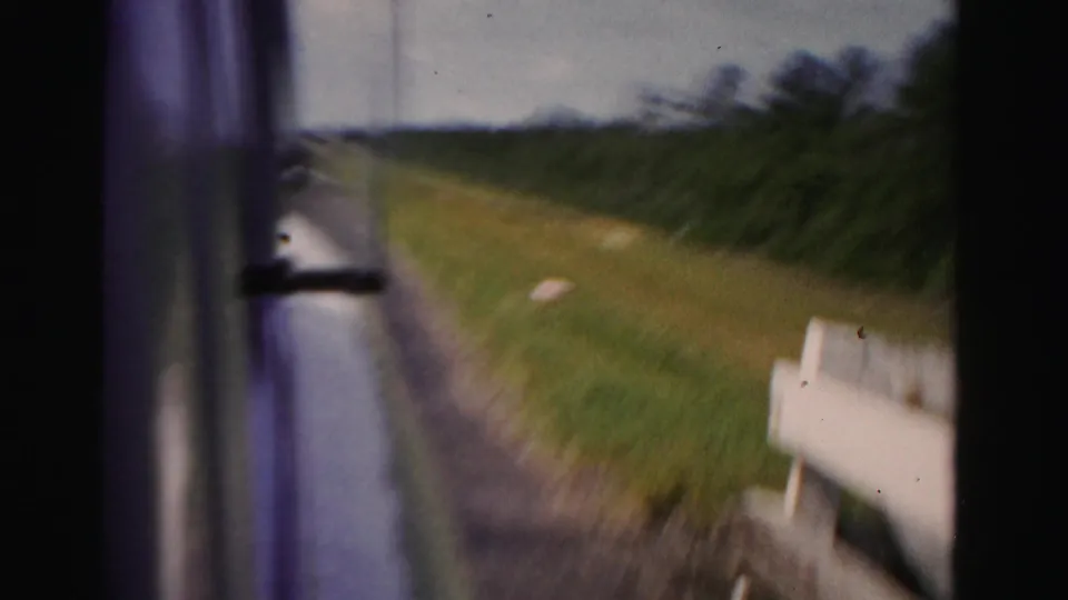 A later frame from the moving car shows the guardrail falling out of view, with grassy roadside and trees in Dade County, Florida.