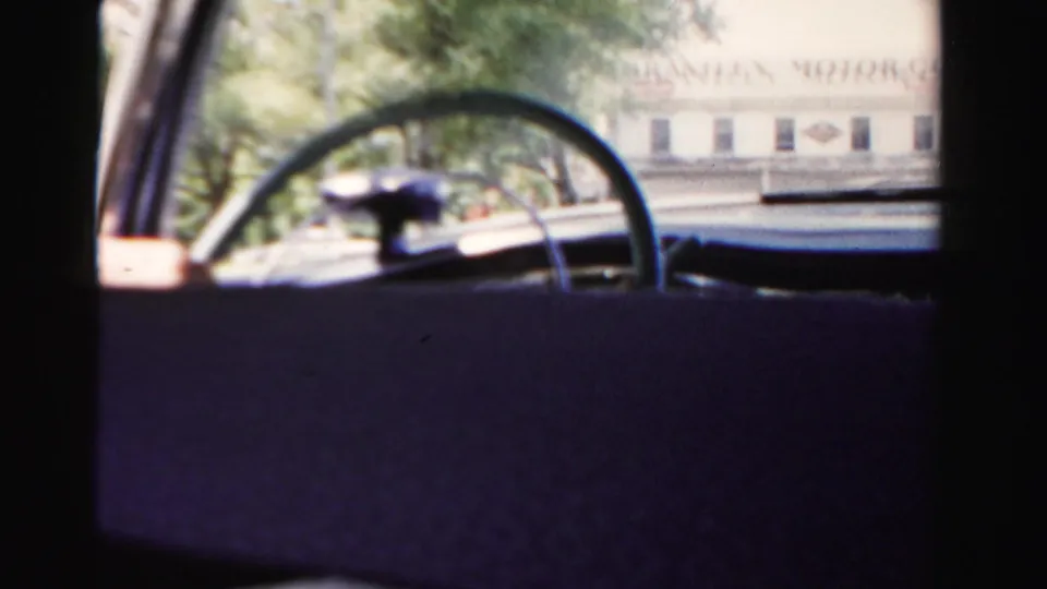 Passenger view across a 1950s car interior toward the steering wheel and a sunlit roadside building outside.