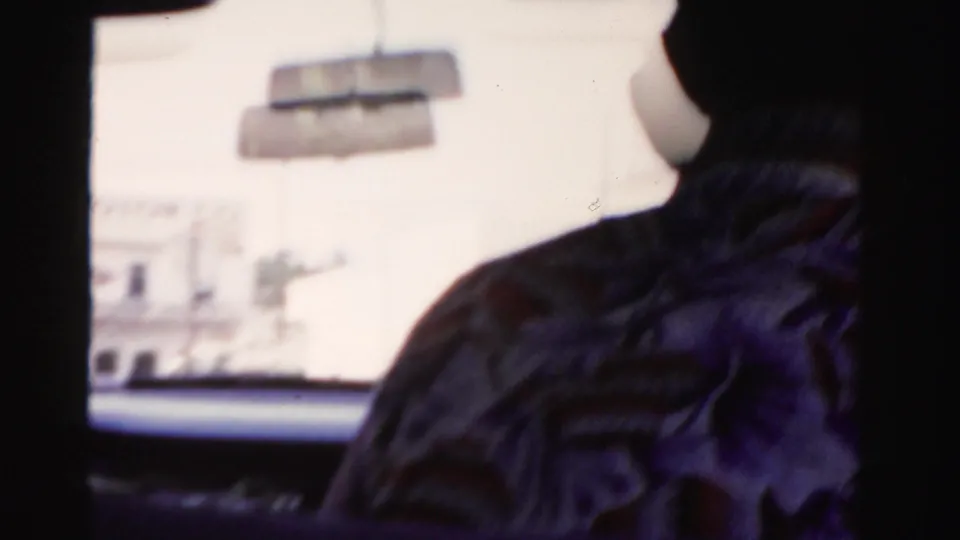 A rear passenger sits in shadow as the car moves past a bright Dade County street scene in 1955 home-movie footage.