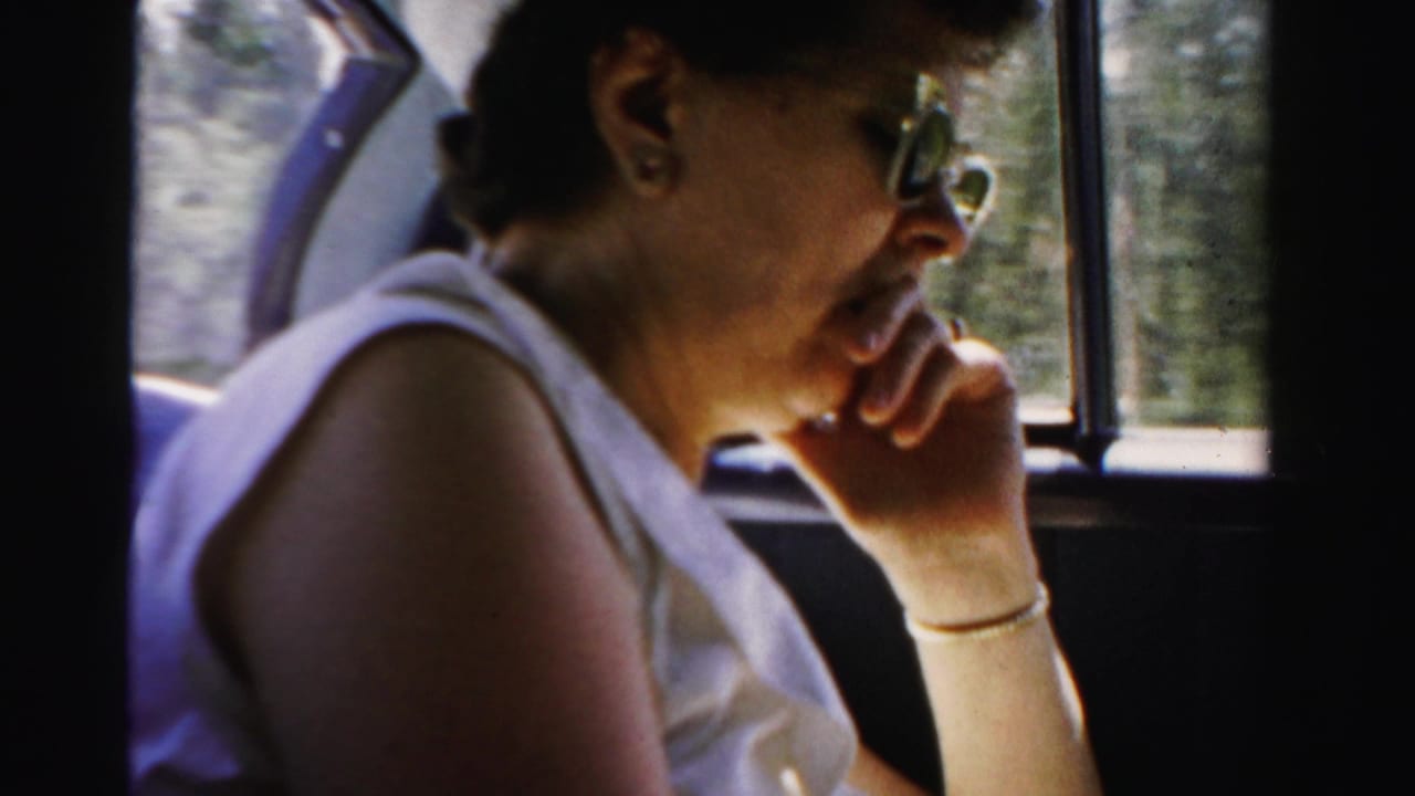 Woman in sunglasses sits by a car window with her hand near her mouth.