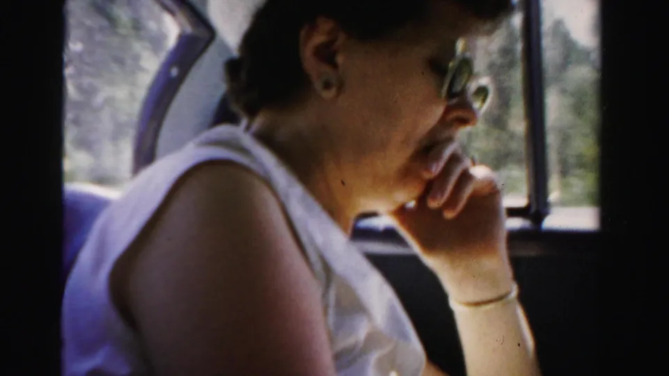 1955 Dade County 8mm frame of a woman in sunglasses, bracelet, and white sleeveless top seated beside a moving car window.