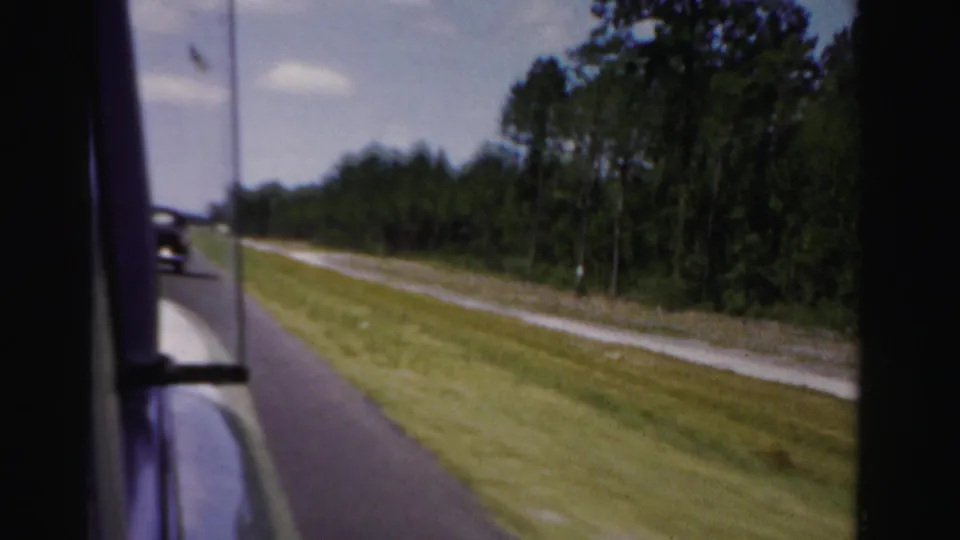 The 8mm view looks from a moving vehicle toward a pine-lined rural road in Dade County, Florida, with a vintage car ahead.