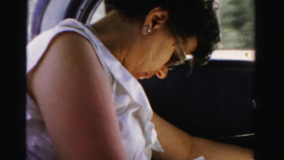 Woman in a white sleeveless top and glasses sits inside the car in 1955 Dade County footage, looking downward as the vehicle travels.