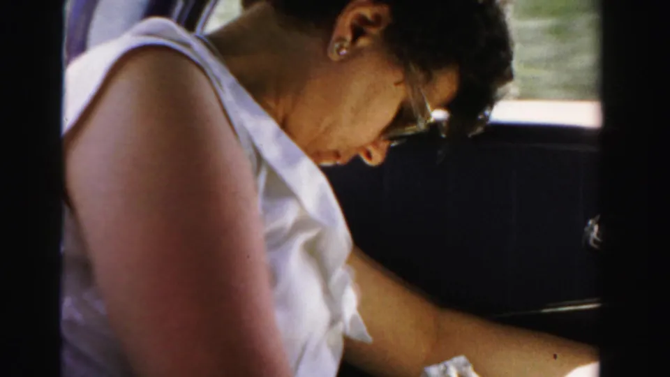 In 1955 Dade County home-movie footage, a woman in glasses rests with her head lowered inside a moving car.