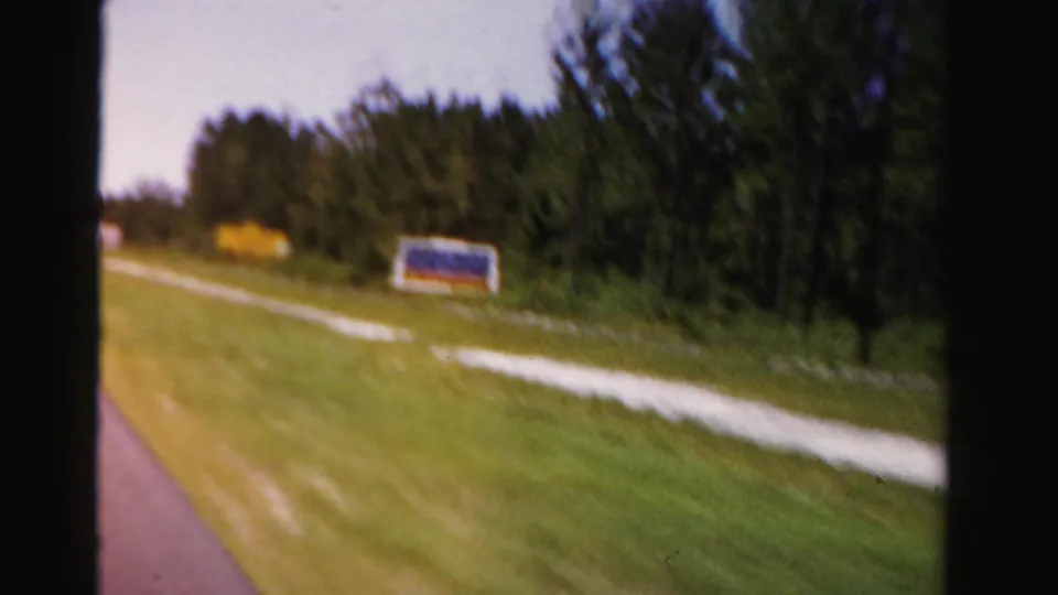 A distant roadside sign stands beside grass and trees in this 1955 Dade County in-car travel view.