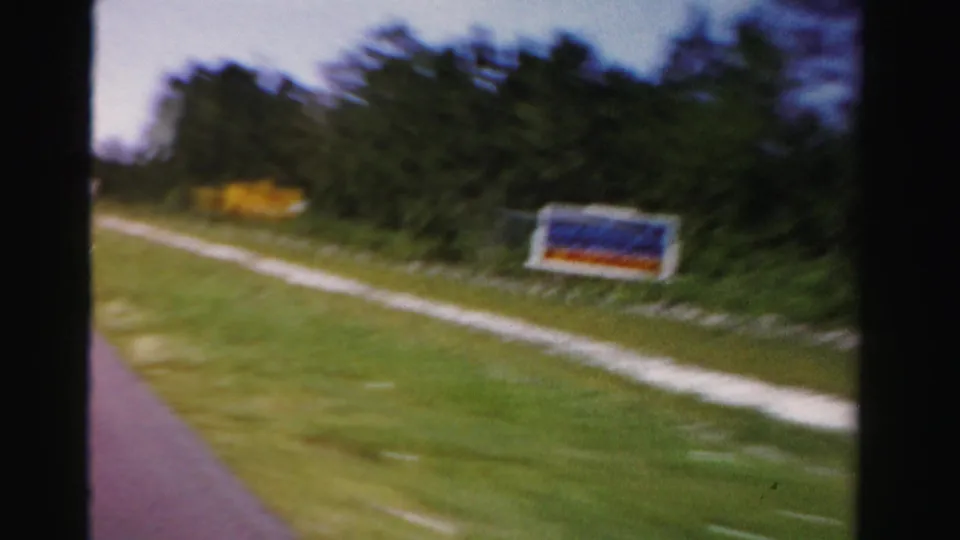 Moving-car view of a roadside sign near a pale dirt path and wooded edge in 1955 Dade County, Florida.