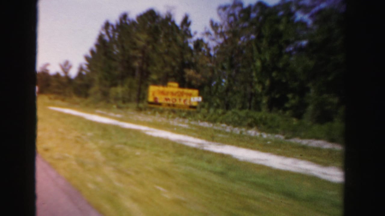 Passing 1955 Florida road view with a yellow sign, tree line, grass, and visible film gate edges.