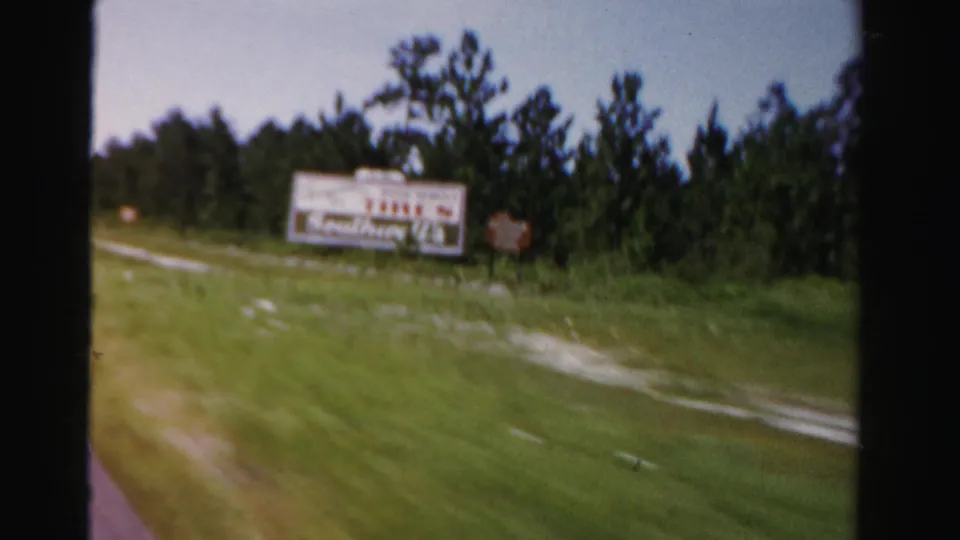 1955 Dade County roadside frame showing a white billboard with red lettering, trees, grass, and car-window framing.