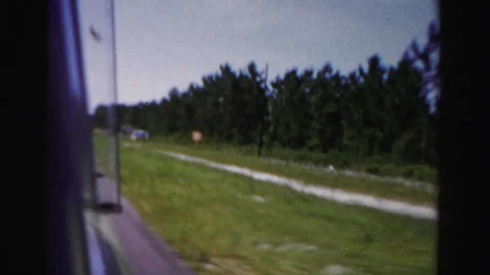 A late-frame 1955 Dade County car-window view shows grass, trees, sky, and small distant roadside signs.
