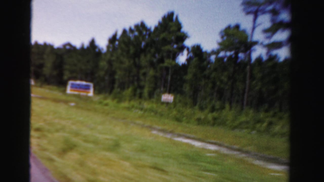 A blue and orange roadside sign passes along a wooded Dade County road in this 1955 car-window view.
