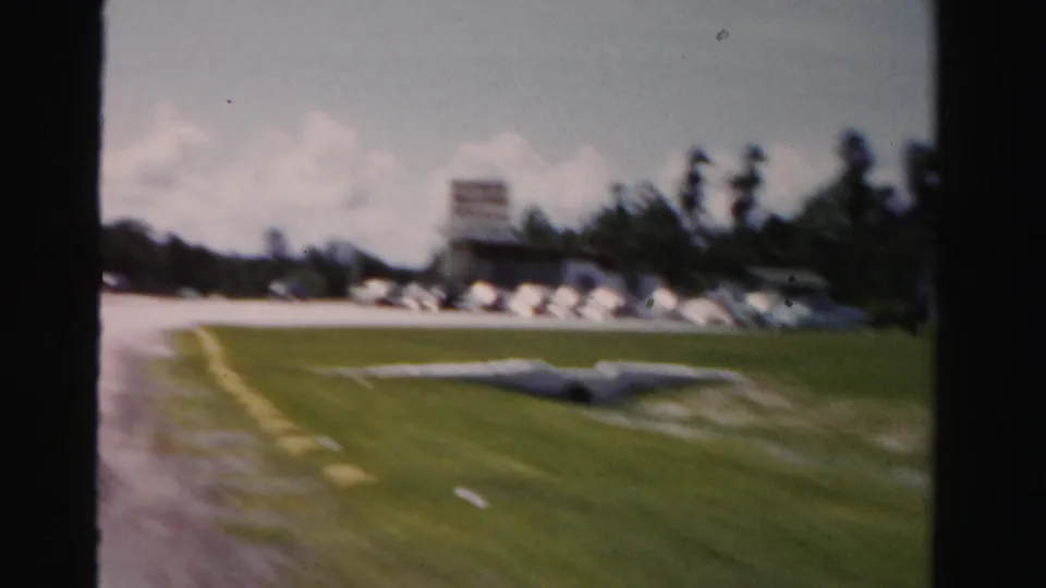 Soft 8mm frame of a curving Dade County roadside with lawn, parked mid-century cars, palms, and a low roadside building.
