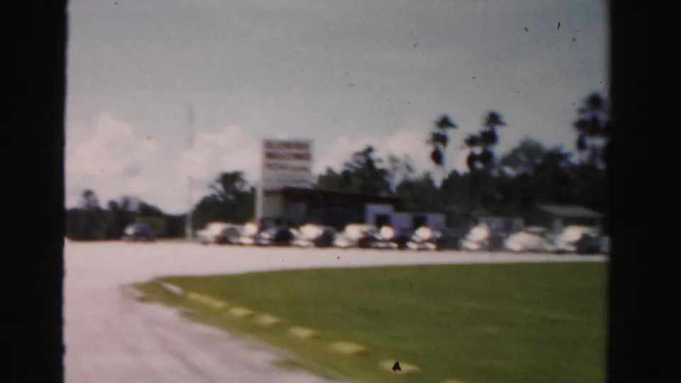 1955 Dade County roadside parking lot with classic cars, a low commercial building, palms, grass, and cloudy sky.