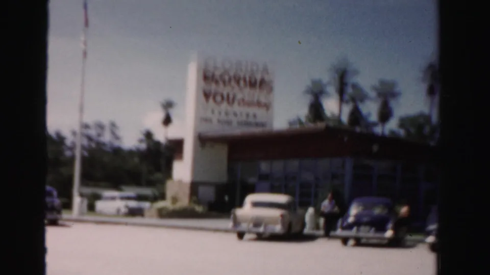 Closer 1955 Dade County roadside view with the welcome sign, mid-century cars, and a few people near the building frontage.