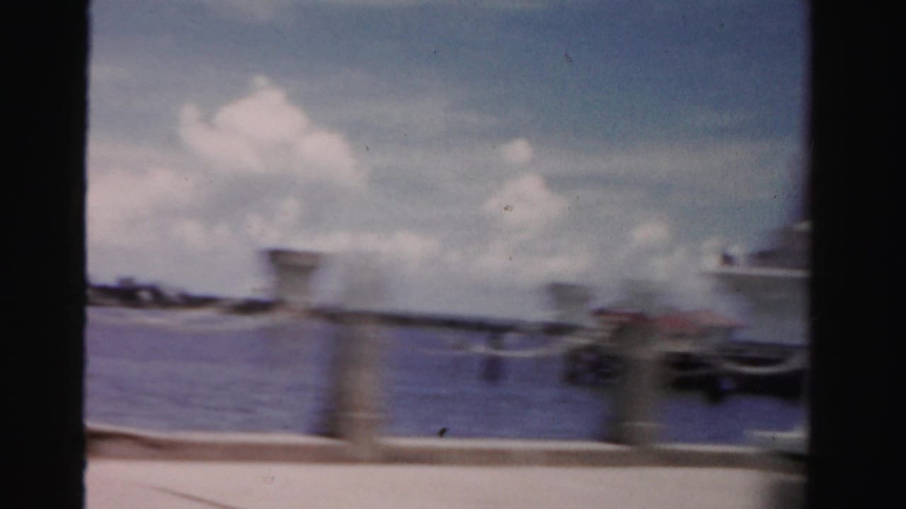 Blurred roadside view from a moving vehicle with water or pavement, sky, posts, and distant cars.