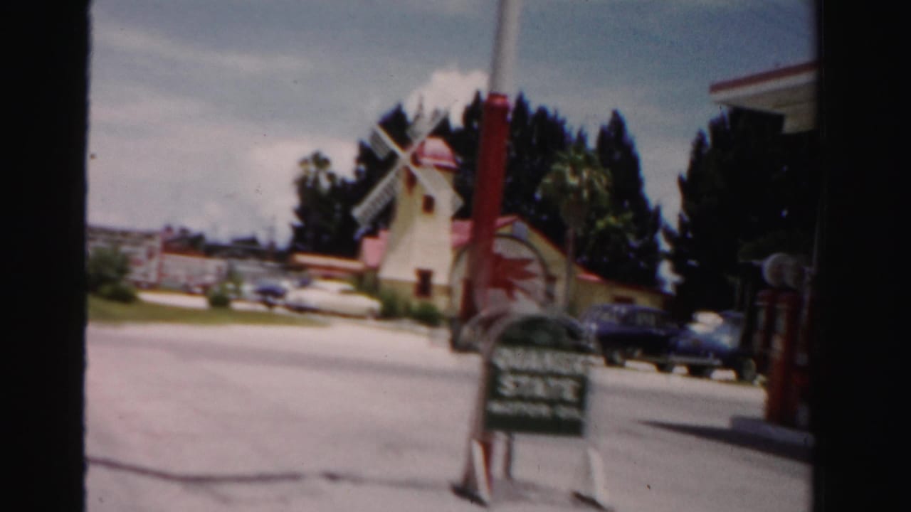 Yellow windmill-shaped building beside a road with a sign, palm trees, and parked cars.