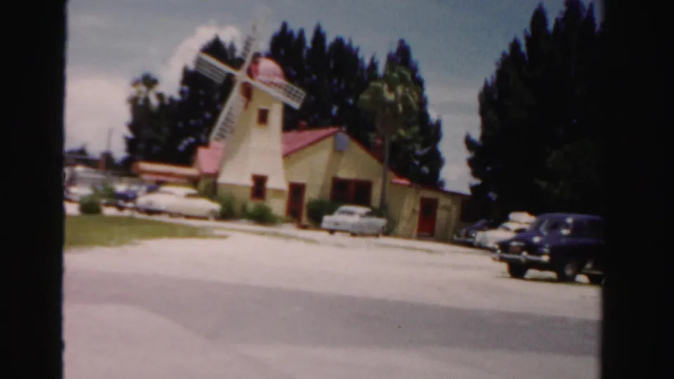 1955 Dade County home-movie frame with the yellow windmill building, red roof, white blades, parked 1950s cars, and roadside greenery.