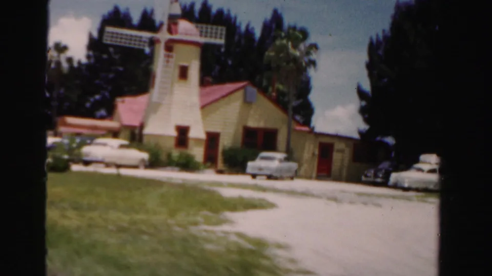 Dade County, Florida 1955 frame showing the yellow windmill roadside building framed by grass, trees, parked cars, and sunny sky.